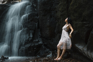 The woman leaned against a moss-covered rock, raising her hands up and touching the rock. The water from the waterfall flows over the rocks next to it, creating a picturesque and natural backdrop.