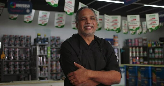 Confident small business owner smiling with arms crossed in his hardware store, shelves of paint cans and supplies in the background, banners hanging from the ceiling