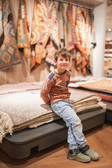Fototapeta premium Smiling boy sitting on folded rugs in a store with colorful patterns in the background.
