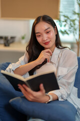 Fototapeta premium Comfortable young asian woman enjoying reading interesting book while sitting on sofa in cozy living room at home, spending leisure time with pleasure
