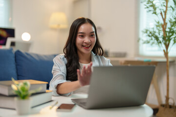 Happy businesswoman waving at her laptop during a video call while working from home, seated at a table surrounded by books and a smartphone in a cozy living room