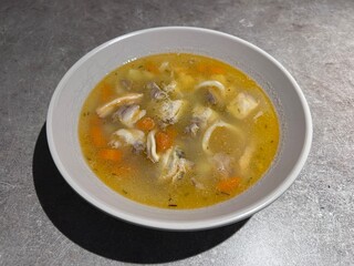 fish soup with vegetables, a spoon on the left side, a yellow napkin under the plate, a white mortar for garlic and small spice jars, on grey background, top view