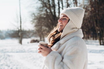 Happy woman with a cup of hot drink in the forest in winter. Winter portrait of a woman in a white...