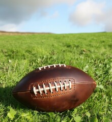 Obraz premium American football resting on green grass field under blue sky with clouds, showcasing leather texture and laces, copy space for text
