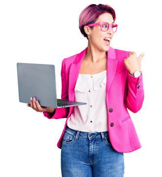 Young beautiful woman with pink hair wearing glasses holding laptop pointing thumb up to the side smiling happy with open mouth