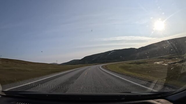 Driving through the tundra in northern Norway on E69 road. Grassy vegetation, coastline, a hill ahead, and the sun create a stunning and tranquil Arctic landscape.