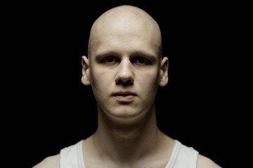 Fototapeta premium Portrait of pale bald man with bridge piercing looking at camera with scary stare standing in dimly lit studio against black background amplifying chilling atmosphere, copy space
