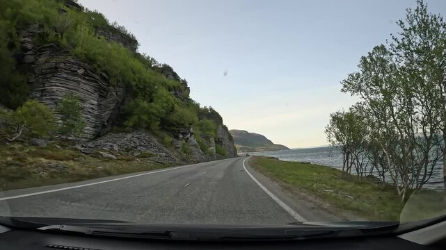 Driving through the tundra in northern Norway on E69 road. Rocky cliffs and low trees line the left side, while the sea stretches on the right. A stunning and remote coastal journey.