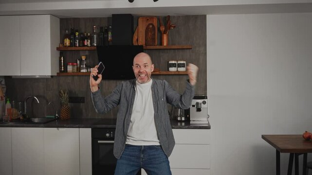 A middle-aged man is in his kitchen when he suddenly receives great news on his smartphone. The expressive man is overjoyed, displaying bright emotions of happiness and excitement.