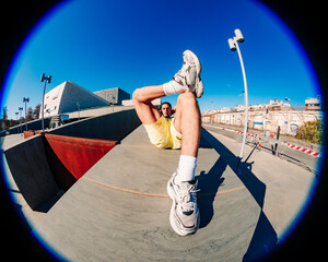 A fisheye view of a man reclining on a modern concrete structure under a vibrant blue sky, emphasizing his sneakers and legs, with urban architecture and a streetlight in the background.
