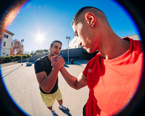 Two young men doing sport activity captured in a fisheye lens perspective, one wearing a red shirt and the other in black, engaging in a handshake under a sunny blue sky