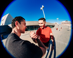 Two young men doing sport activity captured in a fisheye lens perspective, one wearing a red shirt and the other in black, engaging in a handshake under a sunny blue sky