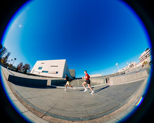 Two men, one in red and one in black, are jogging near a modern urban architectural building under a vibrant blue sky on a sunny day, enjoying the outdoors in a contemporary environment
