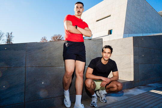 Two male runners one in red and one in black, pose confidently near a modern architectural building under a clear blue sky on a sunny day, exuding style and camaraderie.
