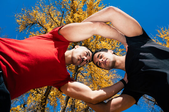 Two male runners, one in a red shirt and one in black, pose together under vibrant yellow autumn leaves, creating a symmetrical and dynamic composition against a backdrop of a clear blue sky.