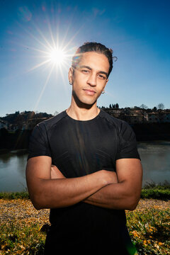 man doing sport in a black shirt standing confidently with arms crossed as the sun creates a starburst effect behind him in a scenic outdoor setting near a calm river under a bright blue sky.
