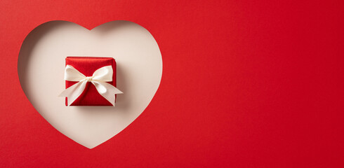 Red gift box with a white ribbon placed inside a paper heart cutout on red. Perfect depiction for Valentines day or romantic holiday themes, emphasizing love, surprise, and special occasions