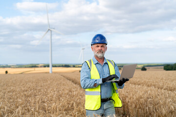 Engineer using laptop in a wheat field with wind turbines in the background during daylight