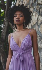 Naklejka premium A striking woman stands gracefully by a stone wall, wearing a flowing lavender dress. Her natural curls frame her face, and she exudes elegance with subtle jewelry.