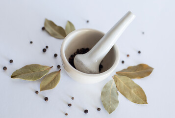 White mortar with black pepper and bay leaves on a white background.
