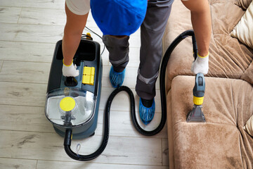 A unique perspective captured from above shows a cleaning company employee in specialized attire, gloves, and shoe covers diligently using a professional vacuum cleaner to clean a sofa.