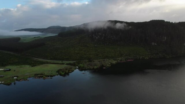 Beautiful aerial view of lake surrounded by forested hills, misty skies, and low clouds.