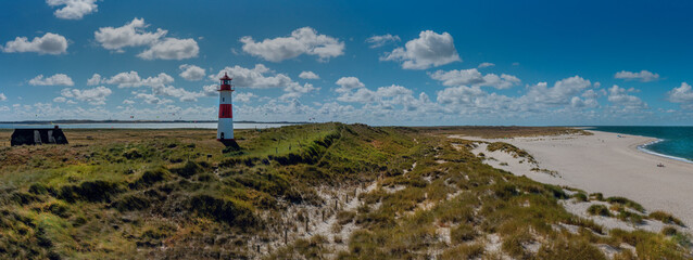 Red-and-white lighthouse on grassy dune, sandy beach, thatched cottage and kite surfers on the horizon at Kite Spot West-List on the Ellenbogen ('Elbow'), Sylt. Panorama of peninsula on northern coast © snapshotfreddy