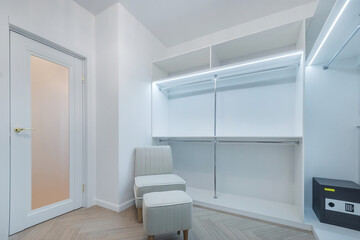 Stylish dressing room with glass entrance door. Cabinets with shelves and a clothes rack. An armchair with an ottoman. Installed safe.