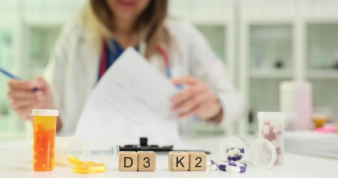 Words D3 and K2 from cubes and drug bottles against doctor writing prescription for patient at workplace in clinic. Woman medic creates recommendations