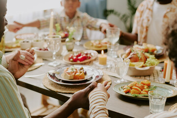 Medium close up of unrecognizable hands of family members holding each other while praying before festive dinner