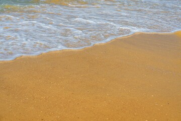 The image showcases a soft wave of the sea gently crashing on the sandy beach, highlighting the wet light brown sand and a small white wave, perfect for beach-themed backgrounds.