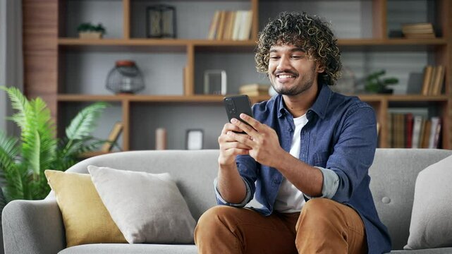 Satisfied happy young man received great news on the mobile phone while sitting on sofa in living room at home. Smiling glad handsome male reads a positive good message on a smartphone, rejoices