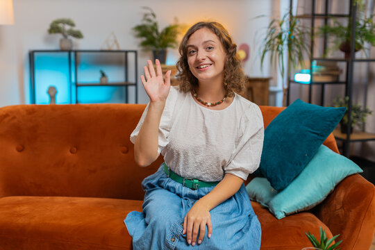Happy young Caucasian woman smiling friendly at camera and waving hands gesturing hello, hi, greeting talking while sitting on sofa in living room at home. Happy girl recording vlog at home. Lifestyle