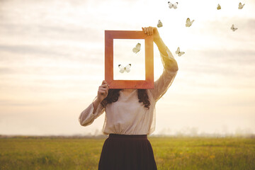 woman holding an empty frame that makes her face disappear reflecting the sky with butterflies...
