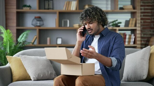 Unhappy young adult man arguing on mobile phone after receiving a parcel with wrong product at home. Angry male makes a bad review, he is dissatisfied with shopping in an online store. Damaged item