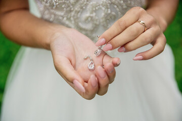 A woman is holding a pair of earrings in her hand. The earrings are silver and have a diamond shape. The woman is wearing a white dress and is standing in a grassy area