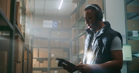 African American Logistics Coordinator in Headset Chats with Customer, Uses Tablet Computer, Looks...
