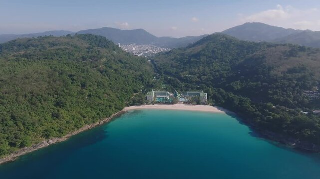 Aerial view of a secluded beach on Phuket Island, Thailand, with turquoise waters, white sand, lush green hills, and a resort near the shoreline.