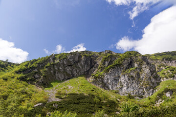 
A steep mountain slope with rocky walls, along which a path winds, but the lower part of the slope is covered with green vegetation.