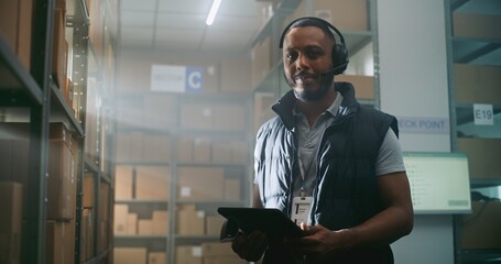 African American Logistics Coordinator in Headset Chats with Customer, Uses Tablet Computer, Looks...