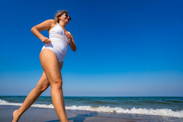 Beautiful middle-aged woman running on sandy beach in summertime. Physical activity on beach. Side view