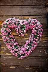 A heart-shaped wreath made of numerous small pink roses hangs on a rustic wooden wall. The contrast between the vibrant flowers and the aged wood creates a visually appealing and charming scene.