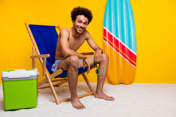 Young male enjoying summer vacation sitting on a beach chair with bright yellow background, surfboard, and cooler.