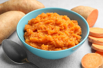 Tasty mashed sweet potato in bowl, spoon and fresh vegetables on gray table, closeup