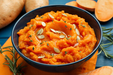 Tasty mashed sweet potato with pumpkin seeds in bowl, fresh vegetables and rosemary on blue wooden table, closeup