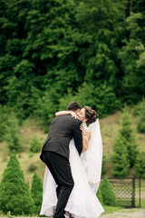 A bride and groom are hugging each other in a lush green forest. The bride is wearing a white dress and the groom is wearing a suit. Scene is happy and romantic