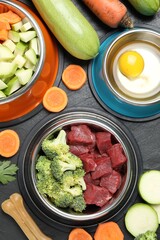 Natural pet food in feeding bowls and products on dark textured table, flat lay