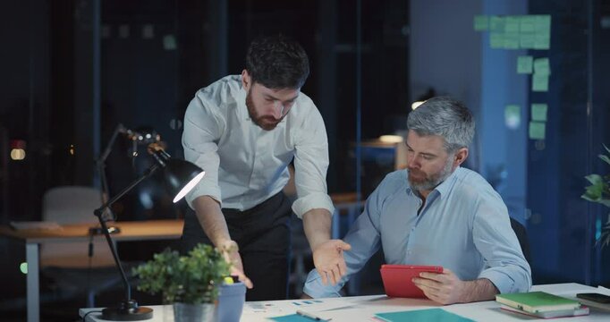 Two caucasian overworked corporate people discussing management ideas soilving financial problem using tablet computer working long hours at office.