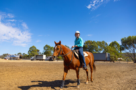 teen girl riding chestnut gelding on bare ground with blue sky and trucks in background