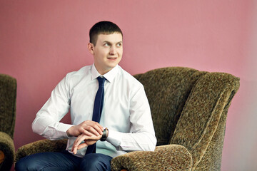 A man in a suit and tie is sitting in a chair with his hands on his hips. He is wearing a watch and...